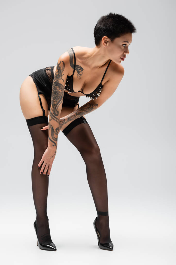 comprimento total de mulher provocante e sexy com o corpo tatuado olhando para longe e posando em sutiã com pérolas, cinto de liga, meias pretas e saltos altos em fundo cinza, arte da sedução  - Foto, Imagem