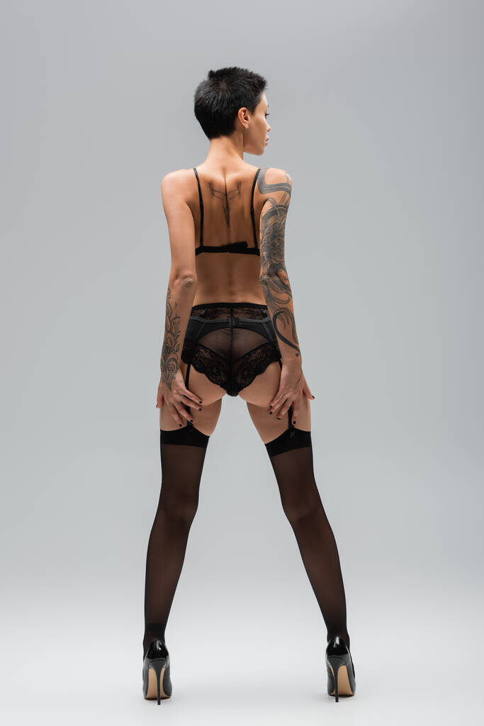 vista posterior de la joven provocativa con pelo corto morena, glúteos sexy y cuerpo tatuado de pie en sujetador negro, bragas de encaje, liguero, medias y tacones altos sobre fondo gris - Foto, Imagen