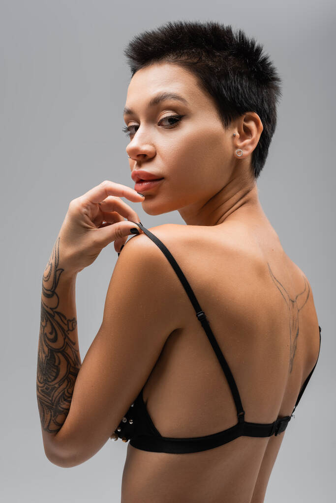 young and flirtatious woman with short brunette hair and sexy tattooed body touching strap of black bra while looking at camera on grey background, erotic photography - Photo, Image