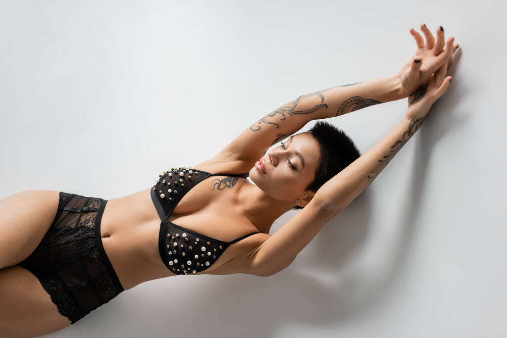 vista superior de la mujer joven, tatuada y deseable con el cuerpo sexy, con sujetador negro con perlas mientras que la colocación en el fondo gris, el arte de la seducción, la fotografía erótica - Foto, imagen