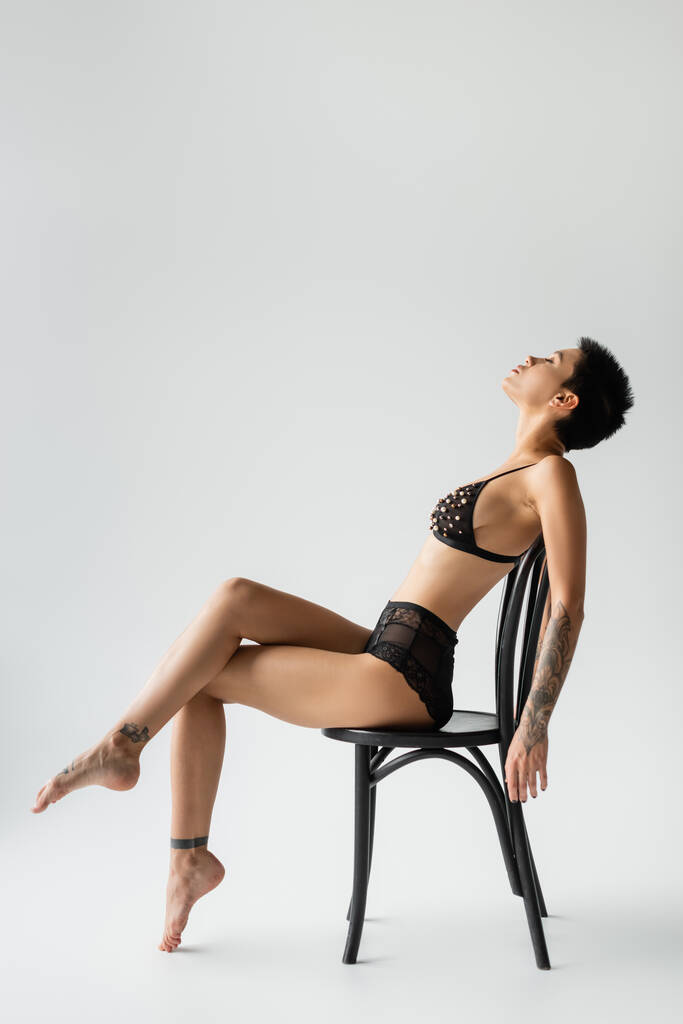 side view of graceful woman with short brunette hair and sexy tattooed body sitting on chair in black bra with pearl beads and lace panties on grey background, erotic photography - Photo, Image