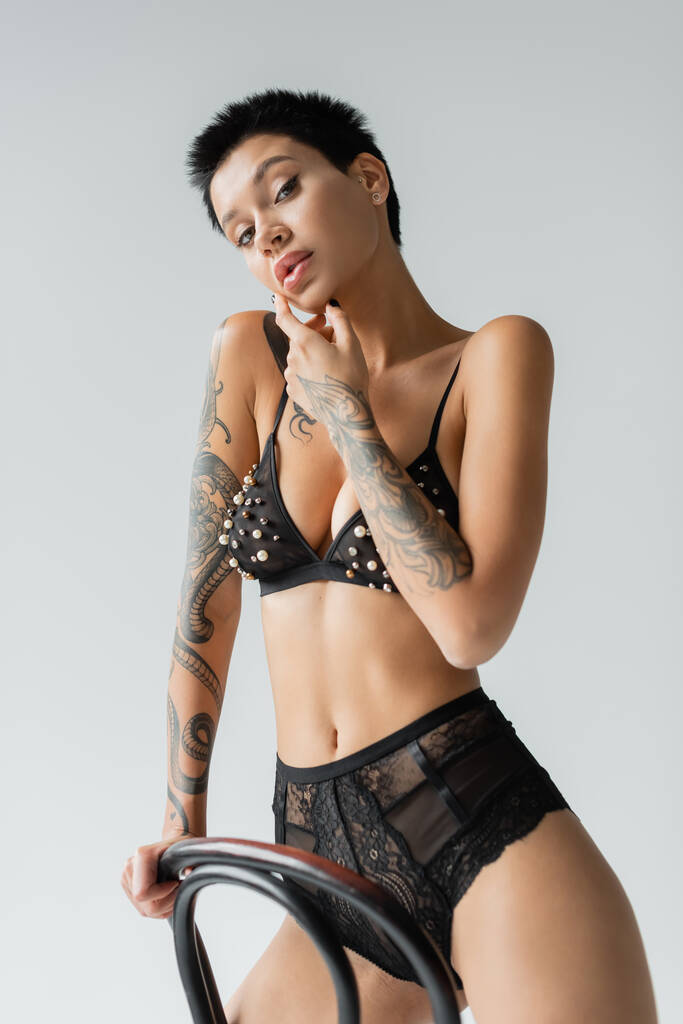 mujer sexy y coqueta con cuerpo tatuado y pelo corto morena, con sujetador negro con cuentas de perlas y bragas de encaje mientras mira la cámara cerca de la silla sobre fondo gris - Foto, imagen