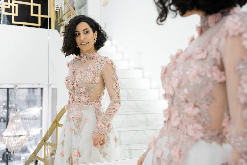брюнетка Среднего Востока женщина с волнистыми волосами, стоящие в великолепном и цветочном свадебном платье, глядя на зеркало в роскошном свадебном салоне, счастливая невеста, очаровательный и элегантный, отражение, покупки - Фото, изображение