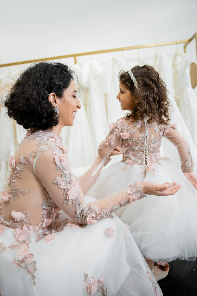 happy middle eastern woman with brunette wavy hair in floral wedding dress looking at tulle skirt of smiling daughter in cute attire in bridal salon, shopping, special moment, togetherness  - Photo, Image