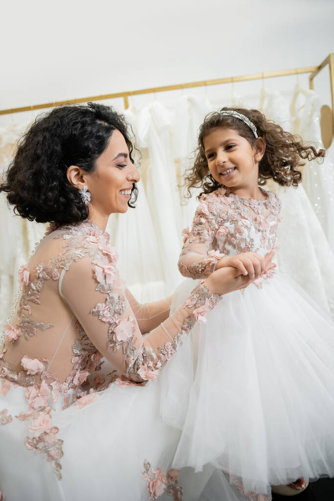 happy middle eastern bride with brunette wavy hair in floral wedding dress holding hands with smiling daughter in cute attire with tulle skirt in bridal salon, shopping, special moment  - Photo, Image