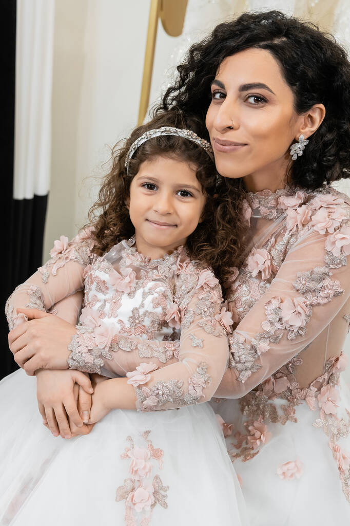 χαρούμενη νύφη της Μέσης Ανατολής με νυφικό αγκάλιασμα χαρούμενο κορίτσι σε χαριτωμένο floral ενδυμασία σε νυφικό σαλόνι, ψώνια, ιδιαίτερη στιγμή, μητέρα και κόρη, ευτυχία, κοιτάζοντας κάμερα - Φωτογραφία, εικόνα