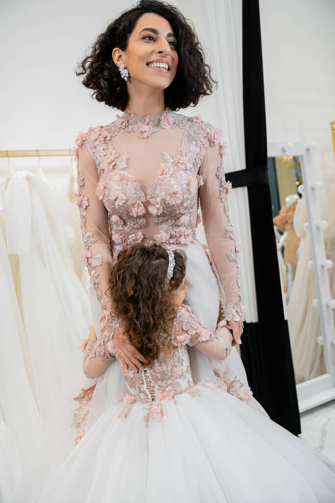 roztomilý dívka v květinovém oblečení objímání veselý matka s brunetkou vlasy stojící ve svatebních šatech v blízkosti rozmazané bílé šaty uvnitř luxusní svatební salon, nakupování, nevěsta-to-be - Fotografie, Obrázek