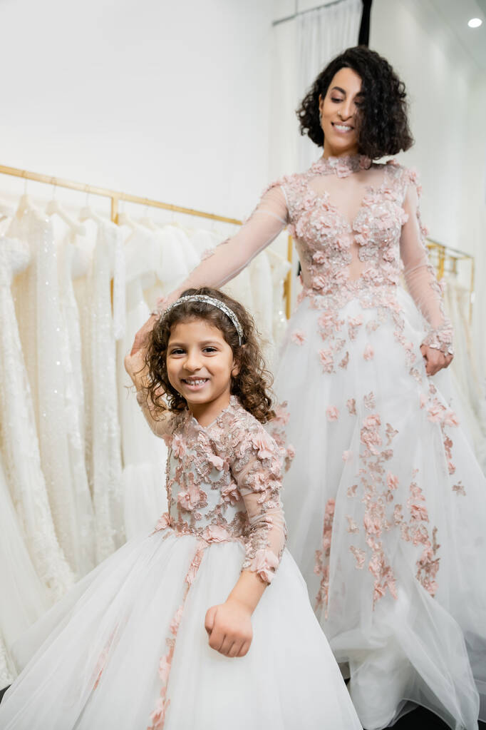 közel-keleti lány virágos öltözékben kéz a kézben boldog nő áll esküvői ruha közelében elmosódott fehér köpeny belsejében luxus menyasszonyi szalon, vásárlás, menyasszony-leendő, anya és lánya  - Fotó, kép