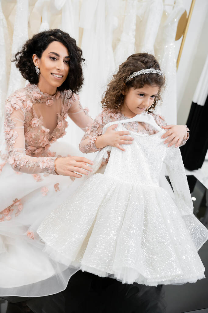 happy and brunette middle eastern bride in floral wedding gown helping to choose dress for her little daughter in bridal salon around white tulle fabrics, process of preparation  - Photo, Image