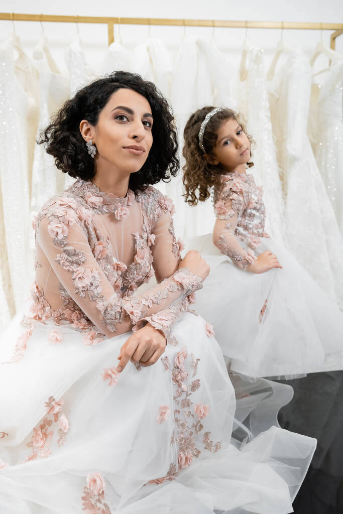 special moment, brunette middle eastern bride in floral wedding gown sitting next to her little daughter in bridal salon around white tulle fabrics, bridal shopping, togetherness  - Photo, Image