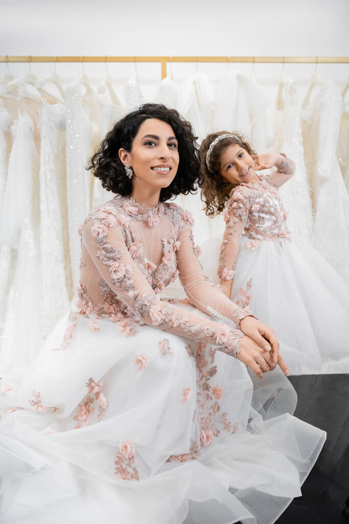 momento especial, encantadora y feliz novia de Oriente Medio en vestido de novia floral sentado junto a su hija pequeña en el salón de novia alrededor de telas de tul blanco, compras nupciales, juntos  - Foto, imagen