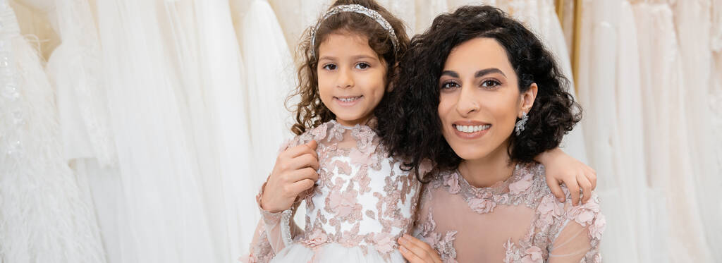 moment spécial, joyeuse mariée du Moyen-Orient en robe de mariée florale assise et embrassant sa petite fille dans un salon de mariée autour de tissus en tulle blanc, shopping nuptial, convivialité, bannière  - Photo, image