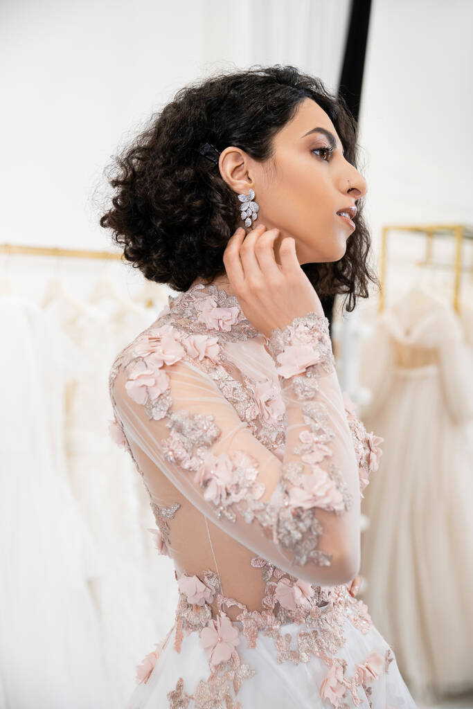 entzückende Frau aus dem Mittleren Osten mit welligem Haar, die in einem wunderschönen und floralen Hochzeitskleid steht und einen anrührenden Ohrring in einem luxuriösen Salon um weiße Tüll-Stoffe trägt, Brauteinkäufe  - Foto, Bild