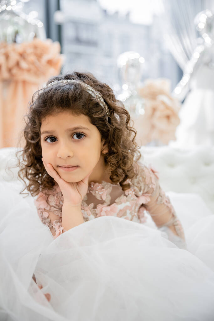 Porträt des netten kleinen Mädchens aus dem Nahen Osten mit lockigem Haar, das in einem floralen Kleid auf einer weißen Couch in einem luxuriösen Hochzeitssalon sitzt, Tüllrock, verschwommener Hintergrund, Blick in die Kamera  - Foto, Bild