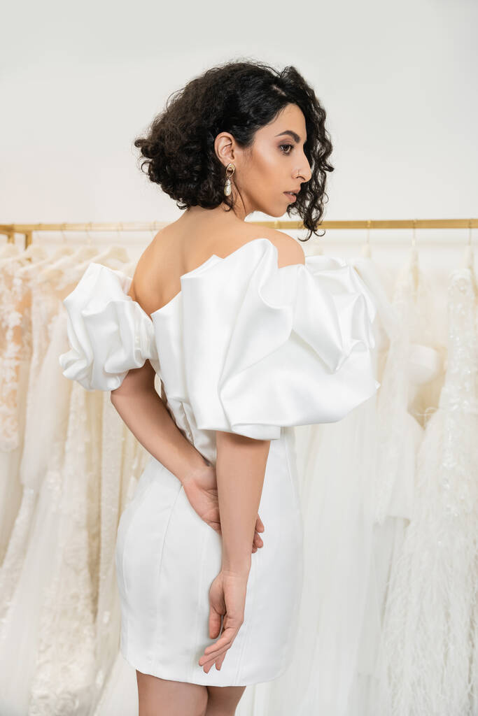 stunning middle eastern bride with brunette and wavy hair posing in stylish wedding dress with puff sleeves and ruffles in bridal boutique next to tulle fabrics, elegant woman  - Photo, Image