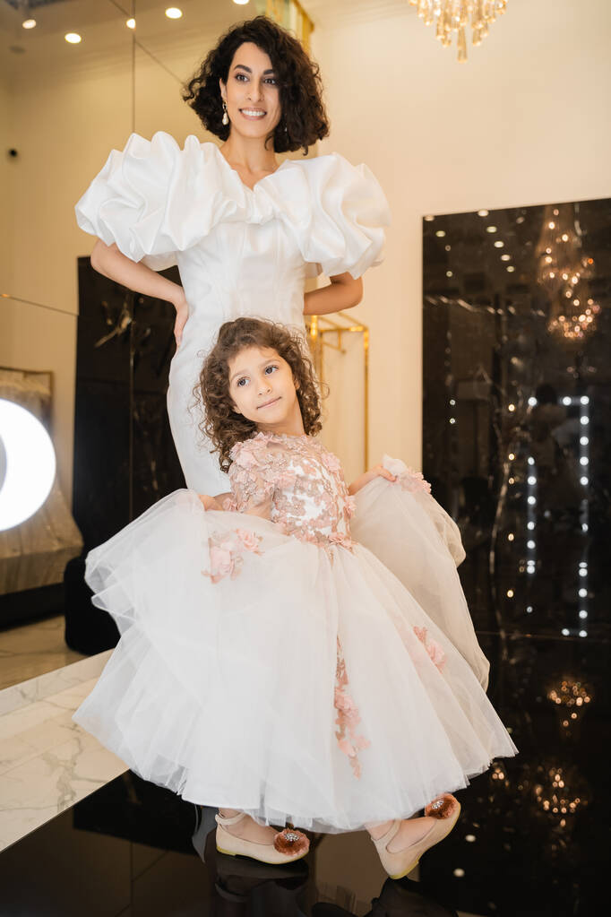 bájos közel-keleti menyasszony barna haj áll fehér esküvői ruha puff ujjak és fodrok és nézi tükör közelében lánya gazdaság tüll szoknya menyasszonyi boltban  - Fotó, kép