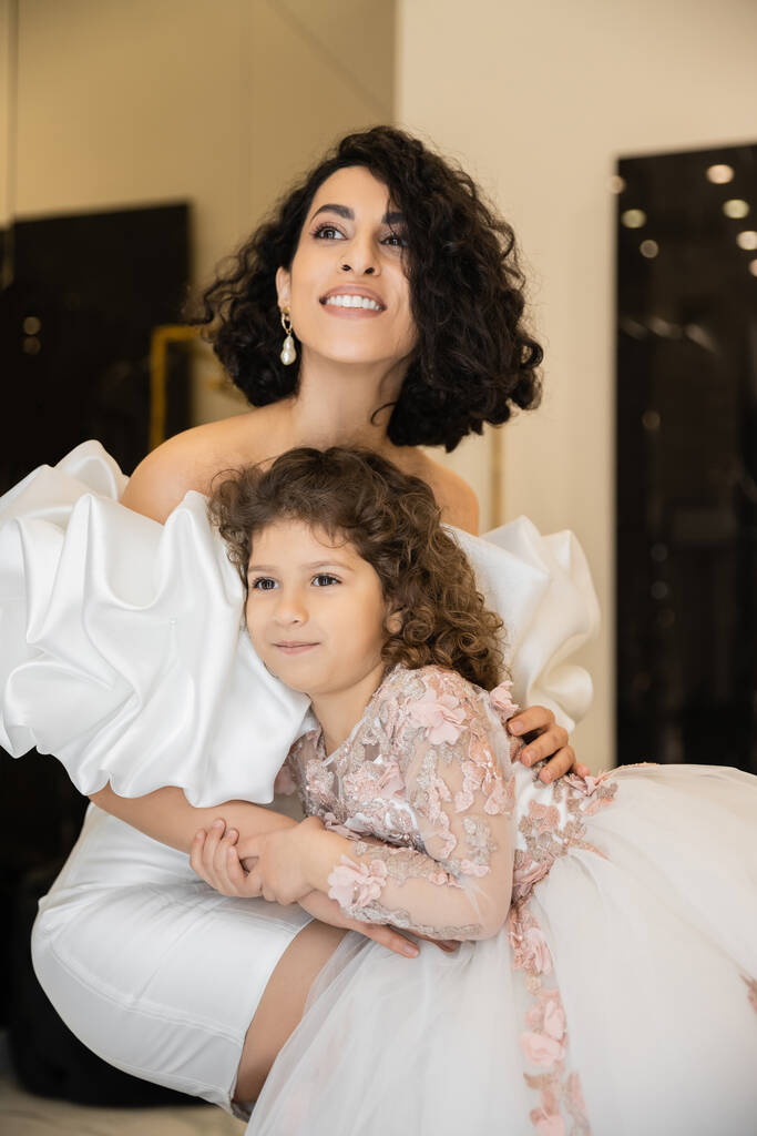 魅力的な中東の花嫁とともにブルネットの髪で白いウェディングドレスでパフスリーブとフリル抱擁の娘と離れてブライダルストア,特別な機会  - 写真・画像