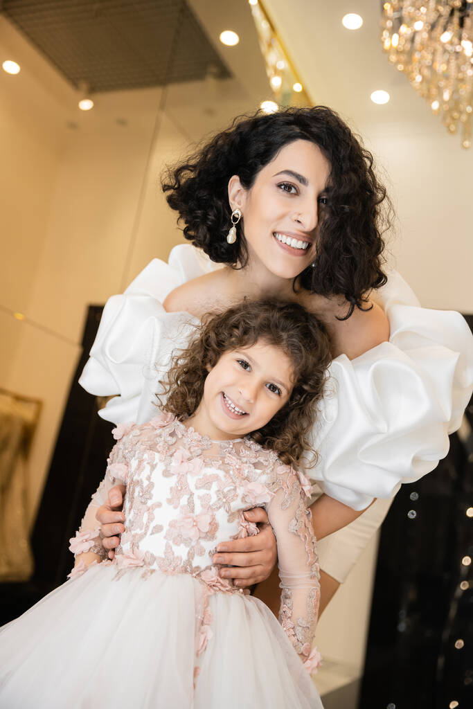 charming middle eastern bride with brunette hair standing in white wedding gown with puff sleeves and ruffles behind cute daughter and smiling together in bridal store, looking at camera  - Photo, Image