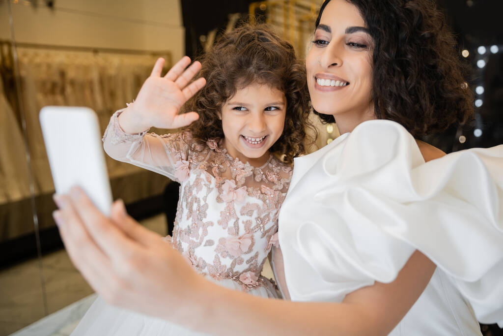 очаровательная невеста с брюнеткой в белом свадебном платье с рукавами и оборками, держащая смартфон, в то время как счастливая дочь машет рукой во время видеозвонка в свадебном магазине  - Фото, изображение