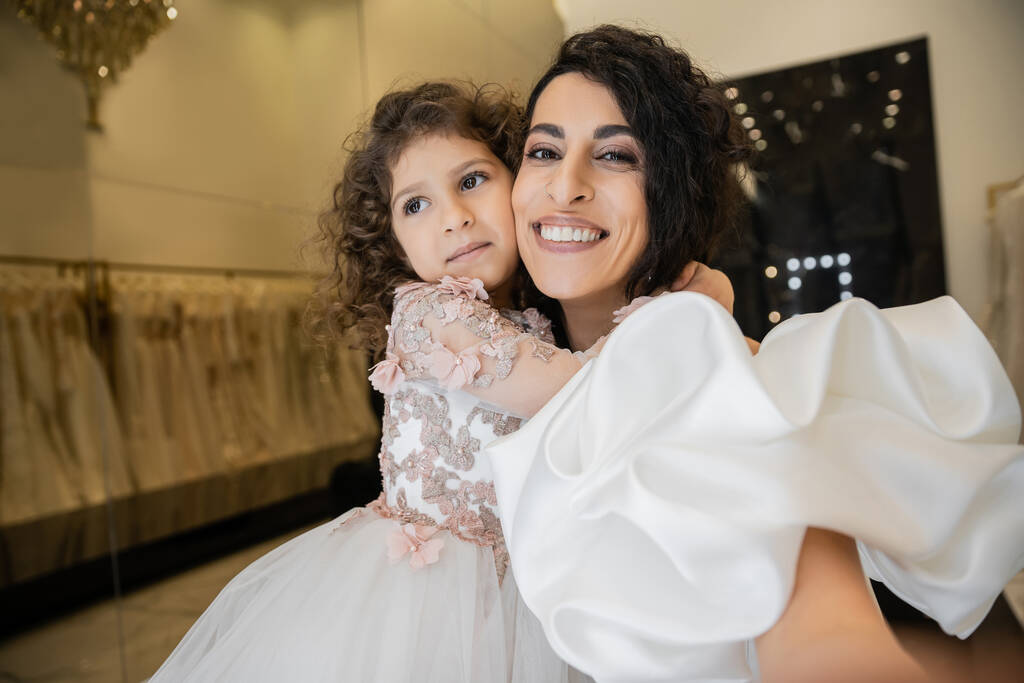 веселая маленькая девочка в цветочном наряде обнимает свою очаровательную мать в белом свадебном платье с пышными рукавами и оборками, улыбаясь и глядя в камеру в свадебном бутике  - Фото, изображение