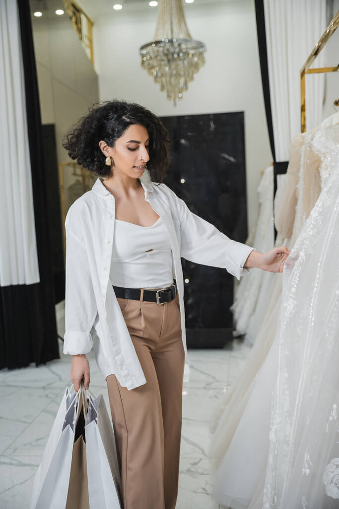 γοητευτική γυναίκα της Μέσης Ανατολής με μελαχρινή και κυματιστά μαλλιά στέκεται σε μπεζ παντελόνι με λευκό πουκάμισο και κρατώντας τσάντες ψώνια, ενώ επιλέγοντας νυφικό φόρεμα στο σαλόνι, ψώνια ξεφάντωμα  - Φωτογραφία, εικόνα