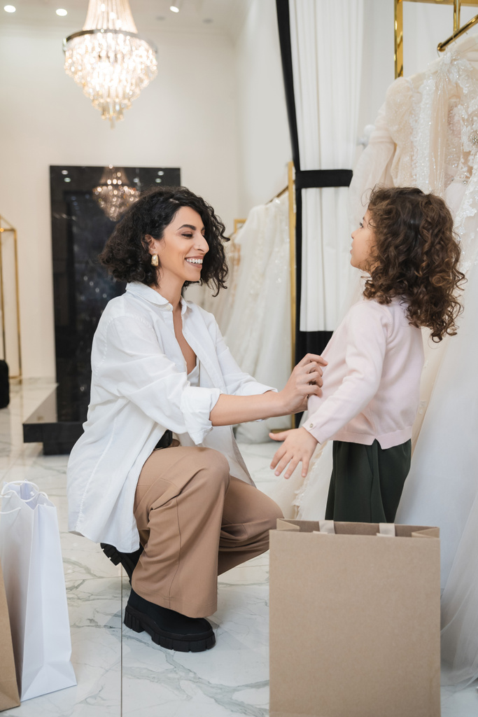 cheerful middle eastern woman with brunette hair in white shirt sitting near shopping bags and wearing cardigan on little girl near wedding dresses in bridal salon, mother and daughter, bride  - Photo, Image
