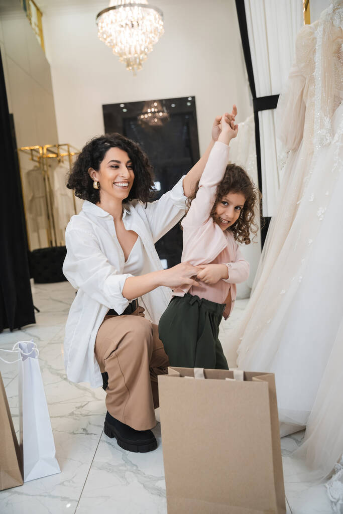 fröhliche Frau aus dem Mittleren Osten mit brünetten Haaren im weißen Hemd sitzt neben Einkaufstüten und hält Händchen von kleinen Mädchen, die neben Brautkleidern im Brautsalon tanzen, Mutter und Tochter, Braut  - Foto, Bild