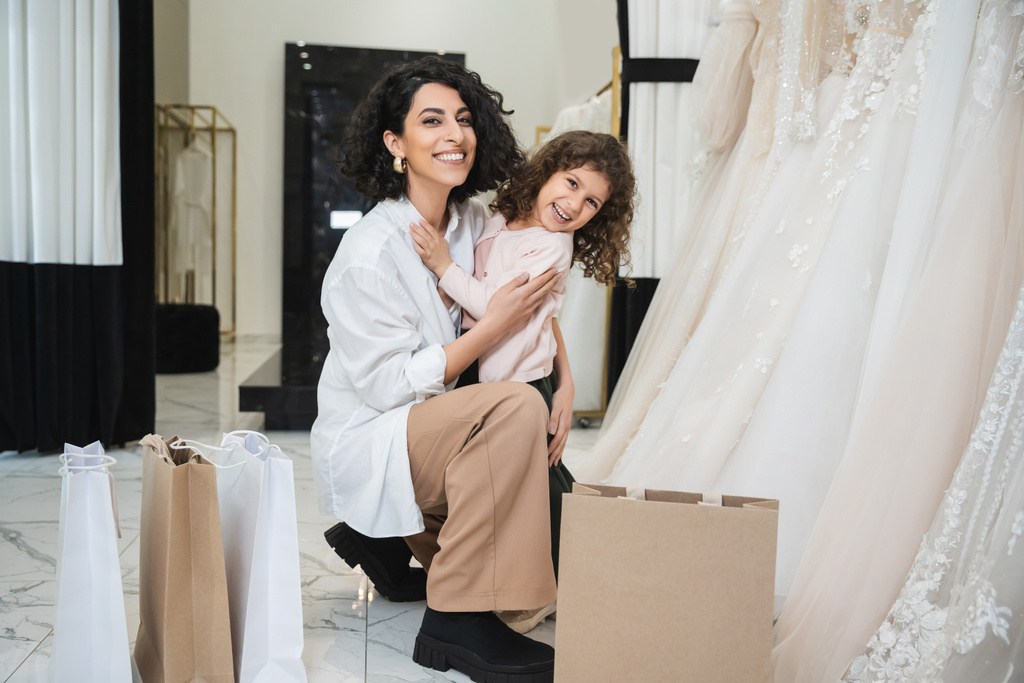 happy little girl hugging cheerful middle eastern bride with brunette hair in white shirt sitting near shopping bags and wedding dresses in bridal salon, mother and daughter, bonding  - Photo, Image