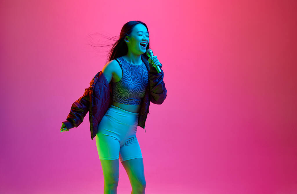 Retrato de niña feliz, sonriente, cantando en micrófono sobre fondo de estudio rosa en luz de neón. Karaoke divertido. Concepto de emociones, expresión facial, juventud, estilo de vida, inspiración, ventas, anuncio - Foto, Imagen
