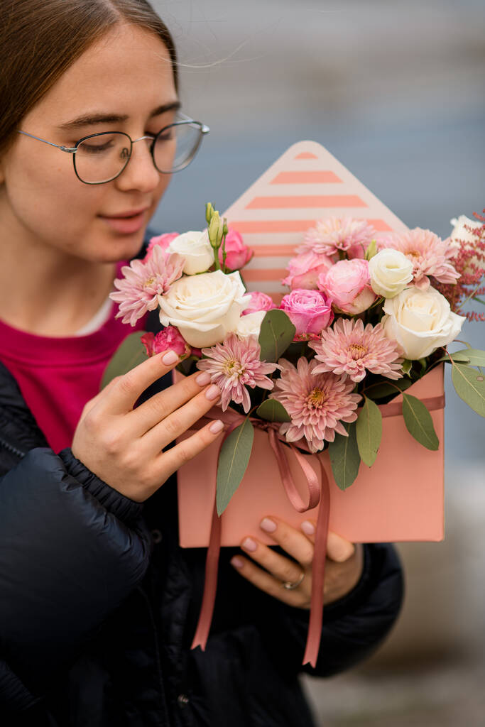 Όμορφη νεαρή γυναίκα κρατά απαλά μπουκέτο με τριαντάφυλλα και χρυσάνθεμα λουλούδια σε ροζ κουτί δώρου με σατέν κορδέλα σε θολή φόντο - Φωτογραφία, εικόνα