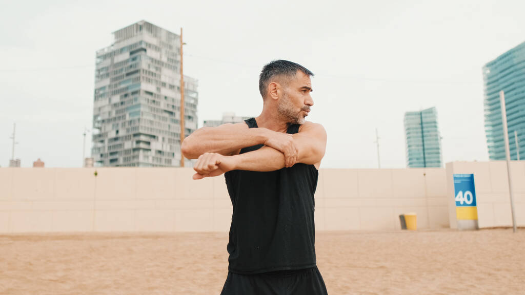 Atlético hombre maduro estira los músculos del brazo mientras está de pie en la playa por la mañana - Foto, imagen