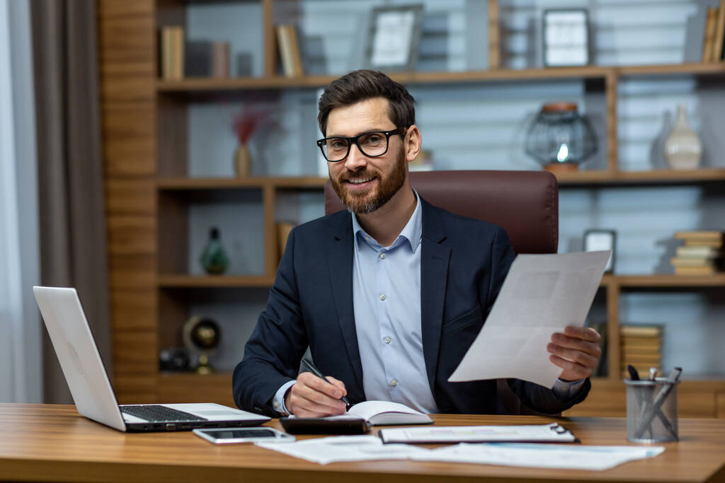 Portret dojrzałego biznesmena w biurze w miejscu pracy, mężczyzna z dokumentami uśmiechnięty i patrzący w obiektyw, papierkowa robota dojrzałego doświadczonego szefa przy biurku. - Zdjęcie, obraz