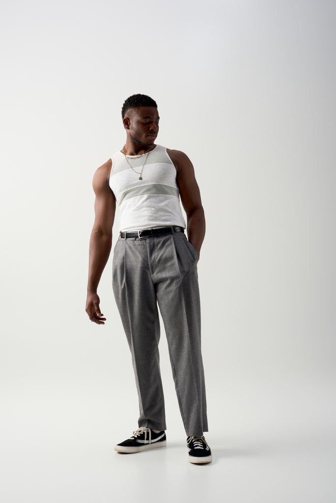 ポケットに手を取りグレーの背景に立つノースリーブのTシャツとパンツを着た若いアフリカ系アメリカ人男性の完全な長さ、スタイリッシュな服装を特徴とする現代的な撮影 - 写真・画像