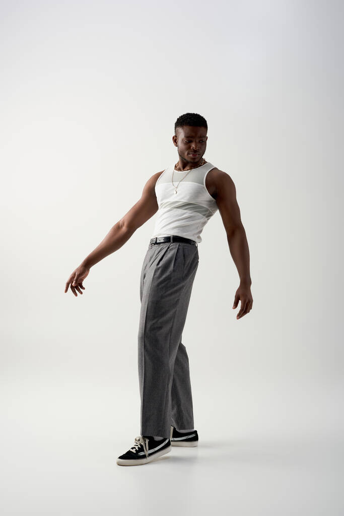 Full length of trendy young african american man in pants, sleeveless t-shirt and sneakers standing on grey background, contemporary shoot featuring casual attire - Photo, Image