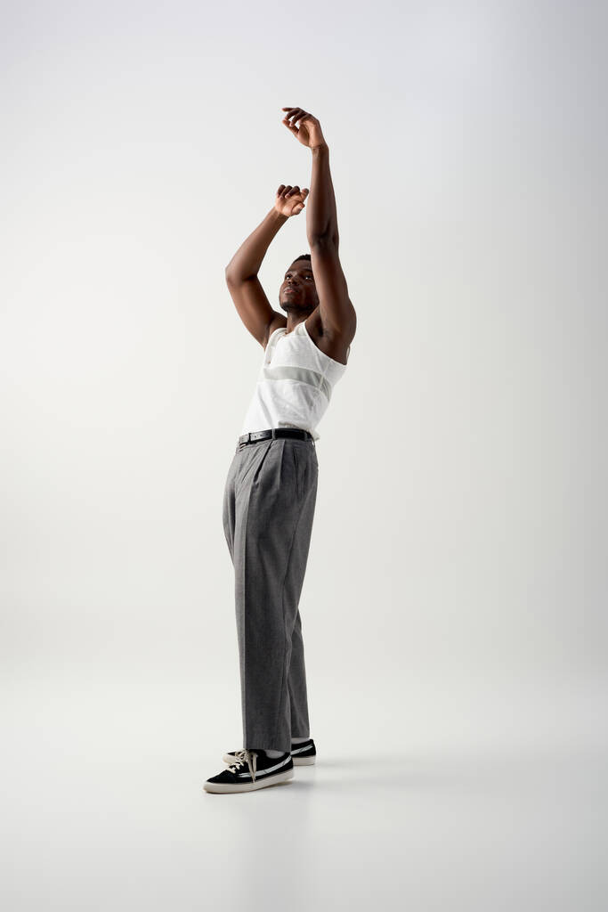 Pleine longueur d'homme afro-américain branché en t-shirt et pantalon sans manches levant les mains et debout sur fond gris, pousse contemporaine avec une tenue élégante - Photo, image