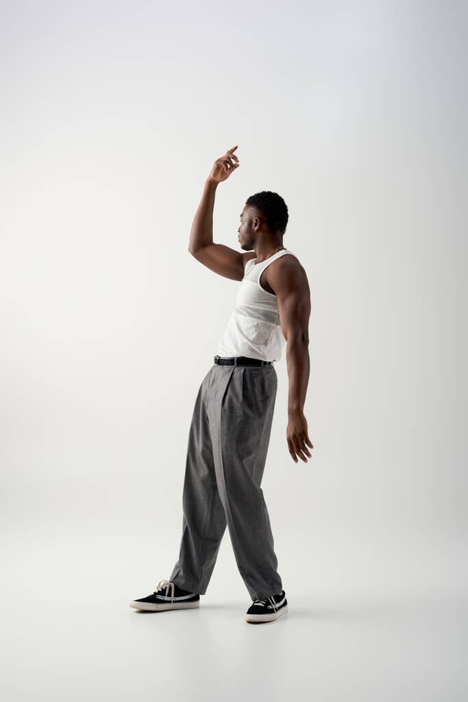 Vista lateral del musculoso joven afroamericano con camiseta sin mangas y pantalones mirando hacia otro lado sobre fondo gris, sesión contemporánea con atuendo casual  - Foto, Imagen