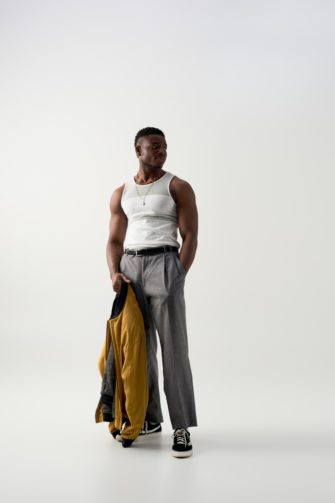 Gri arka planda elinde bomba ceketi olan kolsuz tişörtlü ve pantolonlu modaya uygun genç Afro-Amerikan bir adam. - Fotoğraf, Görsel