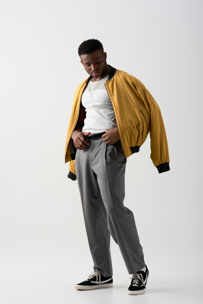Piena lunghezza di fiducioso giovane uomo afroamericano in giacca bomber toccare cintura in vita su pantaloni e in piedi su sfondo grigio, tiro contemporaneo con abbigliamento elegante - Foto, immagini