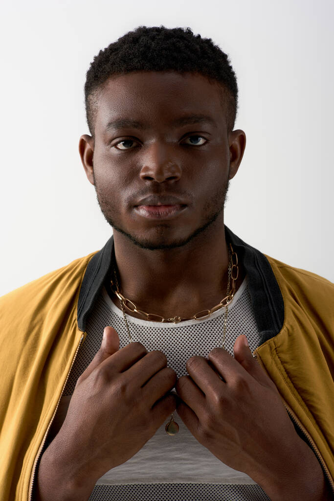 Portrait of young and trendy afroamerican model in bomber jacket and golden necklaces looking at camera isolated on grey, contemporary shoot featuring stylish attire, fashion statement  - Photo, Image
