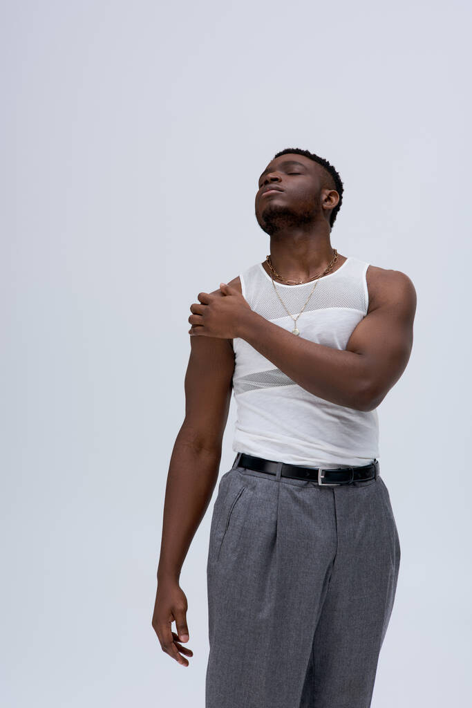 Vue en angle bas du jeune homme afro-américain détendu en t-shirt et pantalon sans manches touchant l'épaule isolé sur gris, pousse contemporaine avec une tenue élégante, musclé - Photo, image