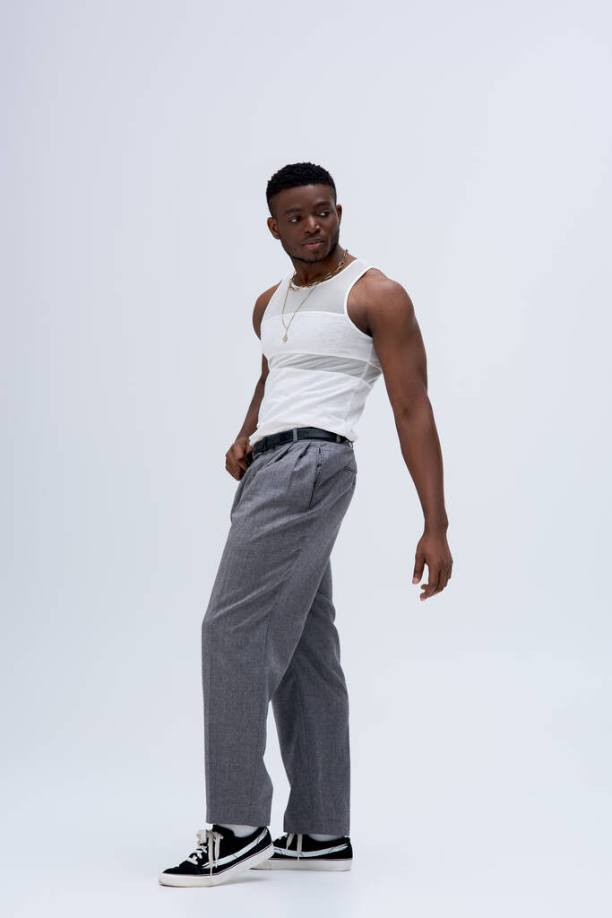 In voller Länge starker afroamerikanischer Mann in Hose und Tank-Top stehend und selbstbewusst posierend in stilvollem und trendigem Outfit auf grauem Hintergrund  - Foto, Bild