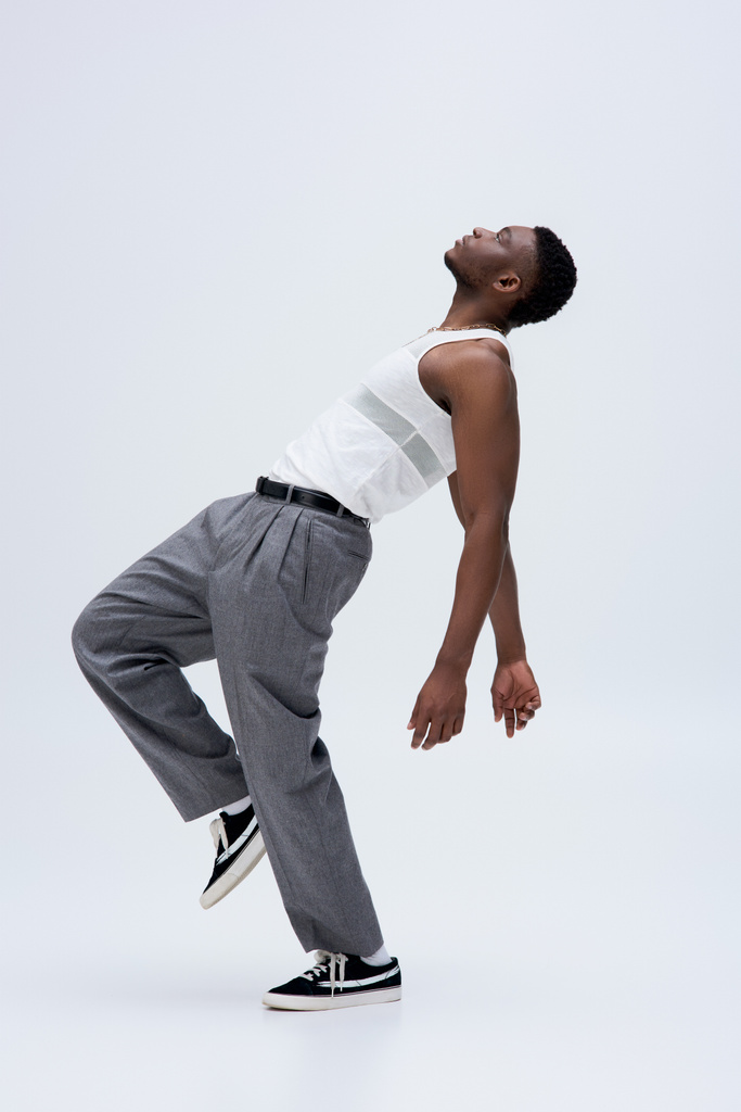 Vista lateral del modelo afroamericano muscular en camiseta sin mangas y pantalones posando con confianza en un atuendo elegante y de moda sobre un fondo gris, atractivo   - Foto, Imagen