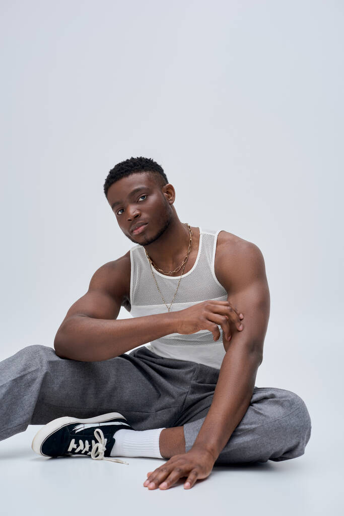 Young african american man in tank top and pants touching arm and posing confidently in stylish and trendy outfit on grey background, good looking, fashion sense  - Photo, Image