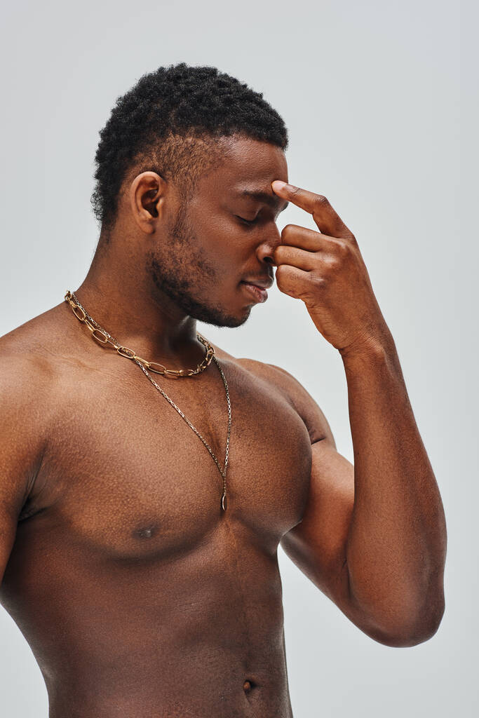 Shirtless and good looking african american model in golden necklaces touching nose isolated on grey, confident and modern pose, fashion shoot - Photo, Image
