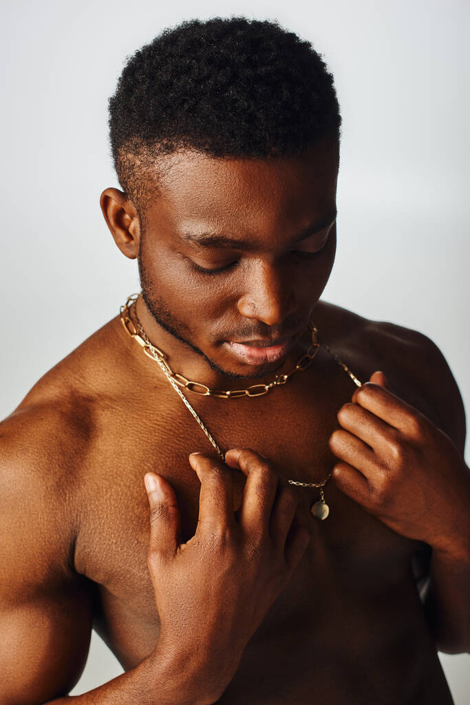 Νεαρό και ημίγυμνο αφροαμερικάνικο ανδρικό μοντέλο που αγγίζει χρυσά περιδέραια και στέκεται απομονωμένο σε γκρι, με αυτοπεποίθηση και μοντέρνα στάση, fashion shoot - Φωτογραφία, εικόνα