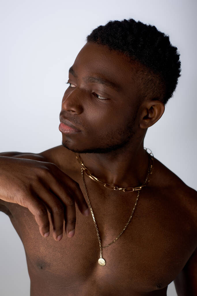 Ritratto di giovane e muscoloso afroamericano in collane dorate che distoglie lo sguardo in piedi isolato su posa grigia, sicura e moderna, servizio fotografico - Foto, immagini