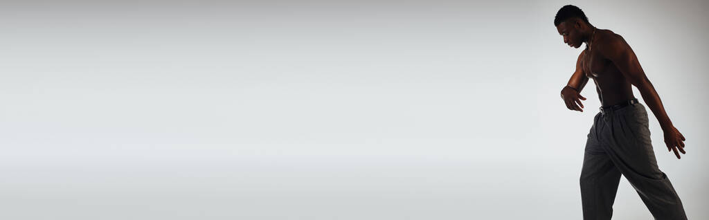 Vista lateral de modelo afroamericano muscular en pantalones y collares de pie en sombra aislada en pose gris, segura y moderna, sesión de moda, pancarta, hombre sin camisa - Foto, imagen