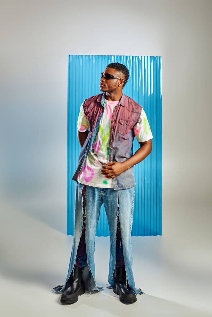 Voller Länge trendiges afrikanisch-amerikanisches Model in farbenfrohem T-Shirt, Sonnenbrille und zerrissenen Jeans, stehend auf grau mit blauem Polycarbonat-Laken im Hintergrund, Mode-Shooting, nachhaltiger Lebensstil - Foto, Bild