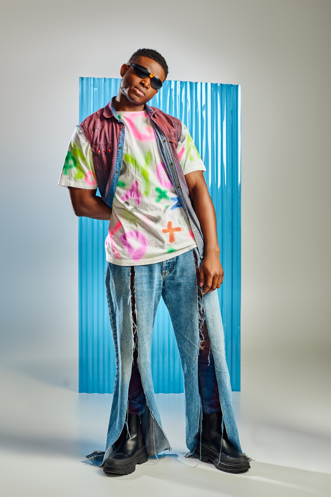 Confident young afroamerican man in colorful t-shirt, sunglasses and ripped jeans standing on grey with blue polycarbonate sheet at background, fashion shoot, sustainable lifestyle - Photo, Image