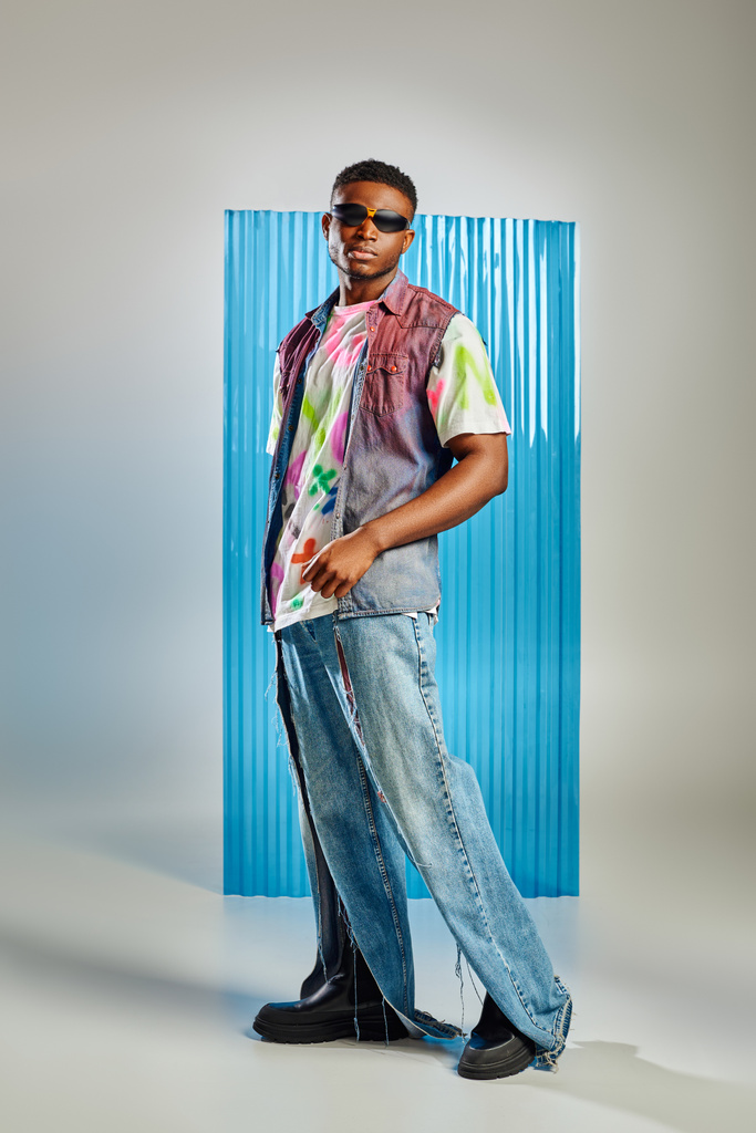 Pełna długość stylowego, młodego modelu afroamerykańskiego w okularach przeciwsłonecznych, podartych dżinsach i dżinsowej kamizelce stojącej na szarym z niebieskim arkuszem poliwęglanu na tle, sesja modowa, odzież DIY - Zdjęcie, obraz